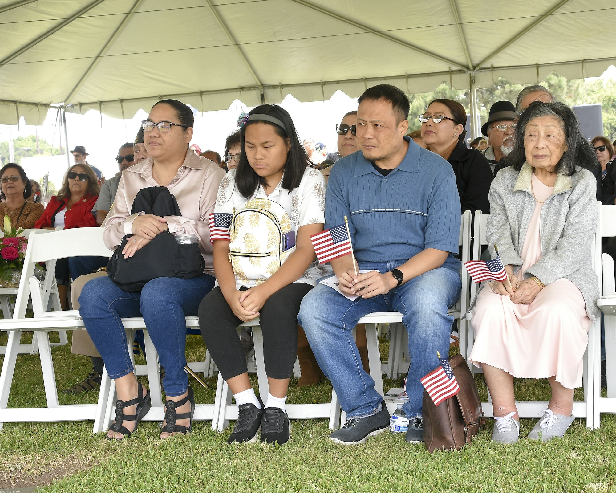 Group of people sitting under a tent, some holding small American flags, at an outdoor event.