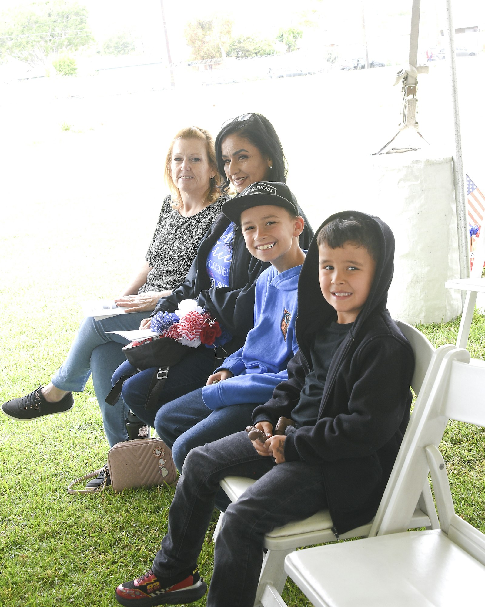 Three people seated outdoors with smiles; two women and a boy wearing a cap. There's a green field in the background.