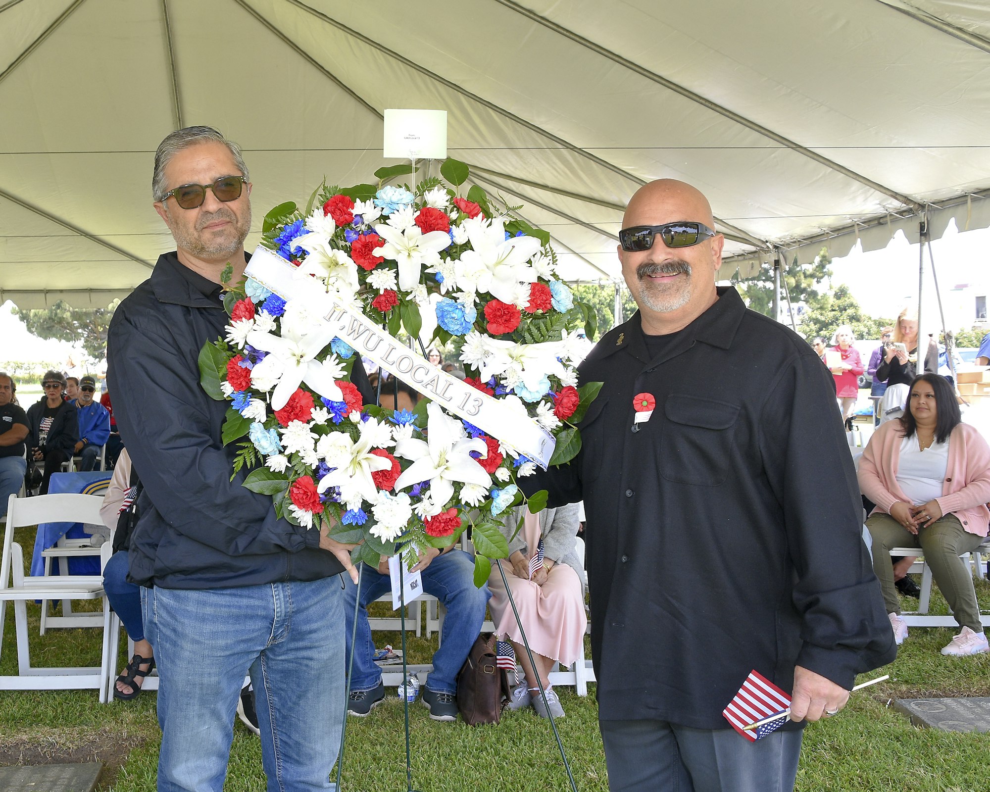 Two men holding a floral wreath with "ILWU LOCAL 13" ribbon, outdoors at an event, possibly a memorial with attendees in the background.