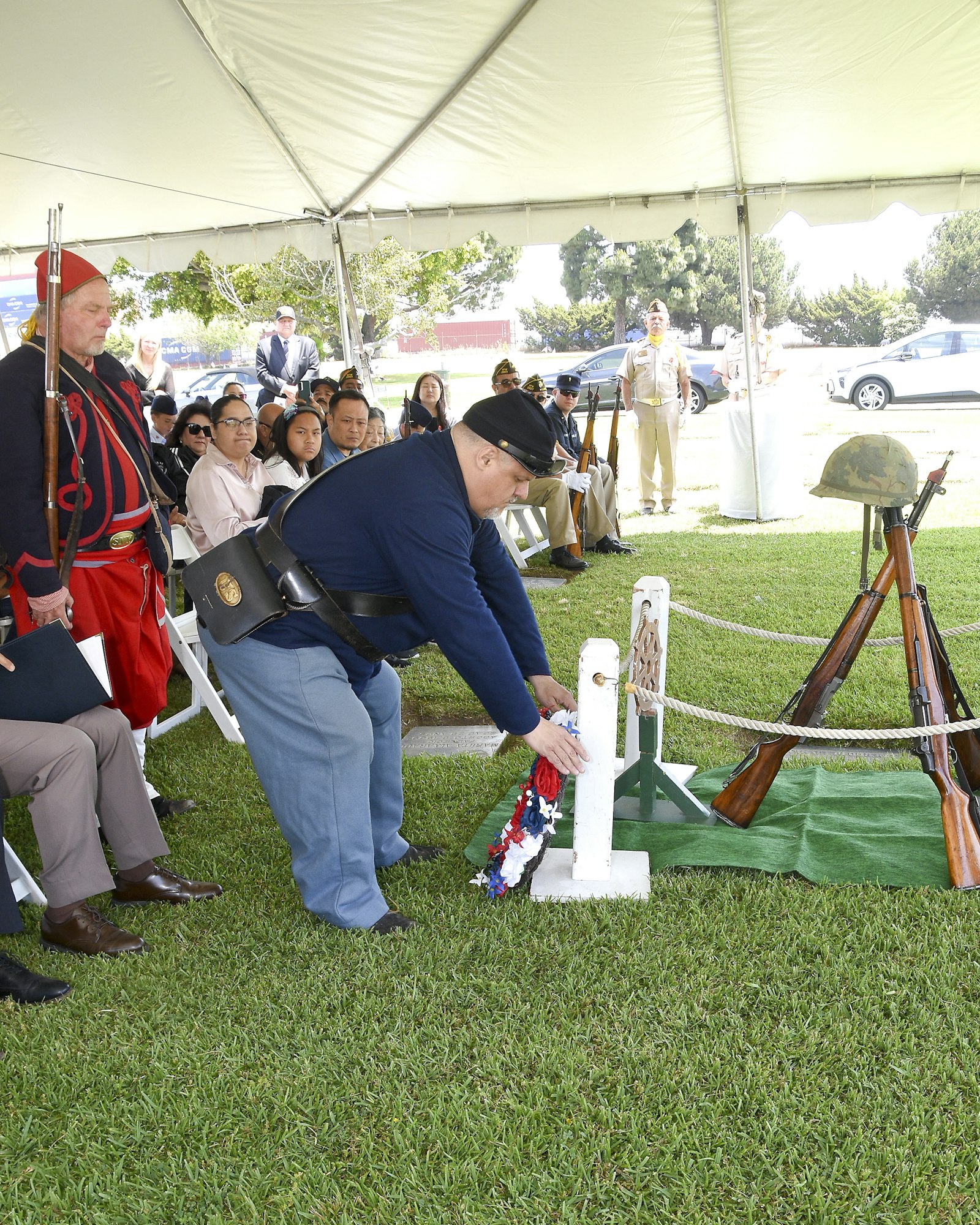 A person in a military uniform placing a wreath at a memorial cross, with onlookers in contemporary and historical military attire.