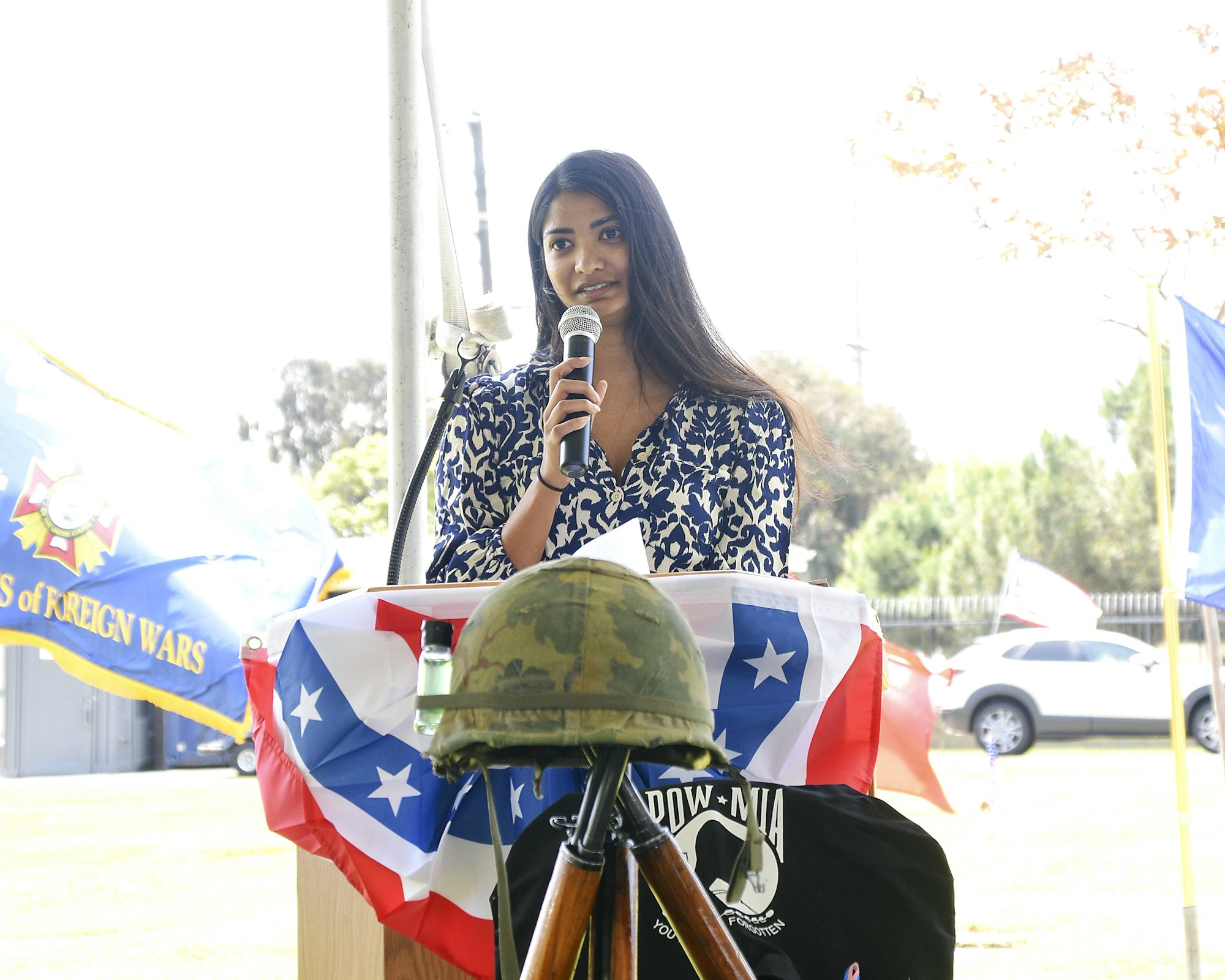 A person speaking into a microphone at an outdoor event with flags and a helmet on the table.