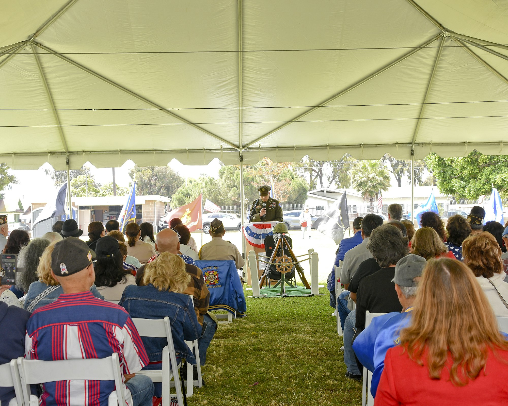 A person in military uniform speaks to an audience under a large tent, with flags in the background.