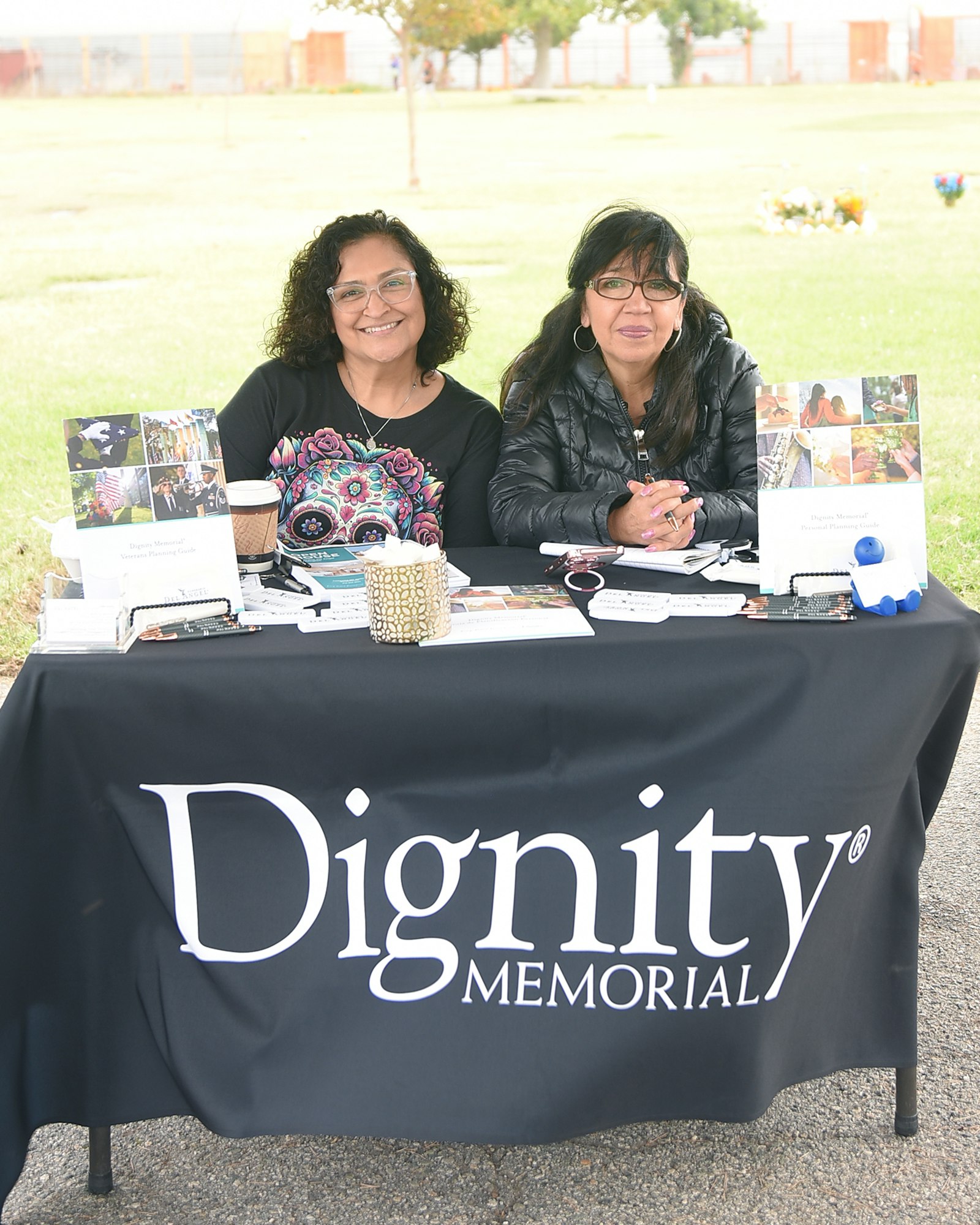 Two people seated behind an information booth with a "Dignity Memorial" banner and promotional materials on the table.