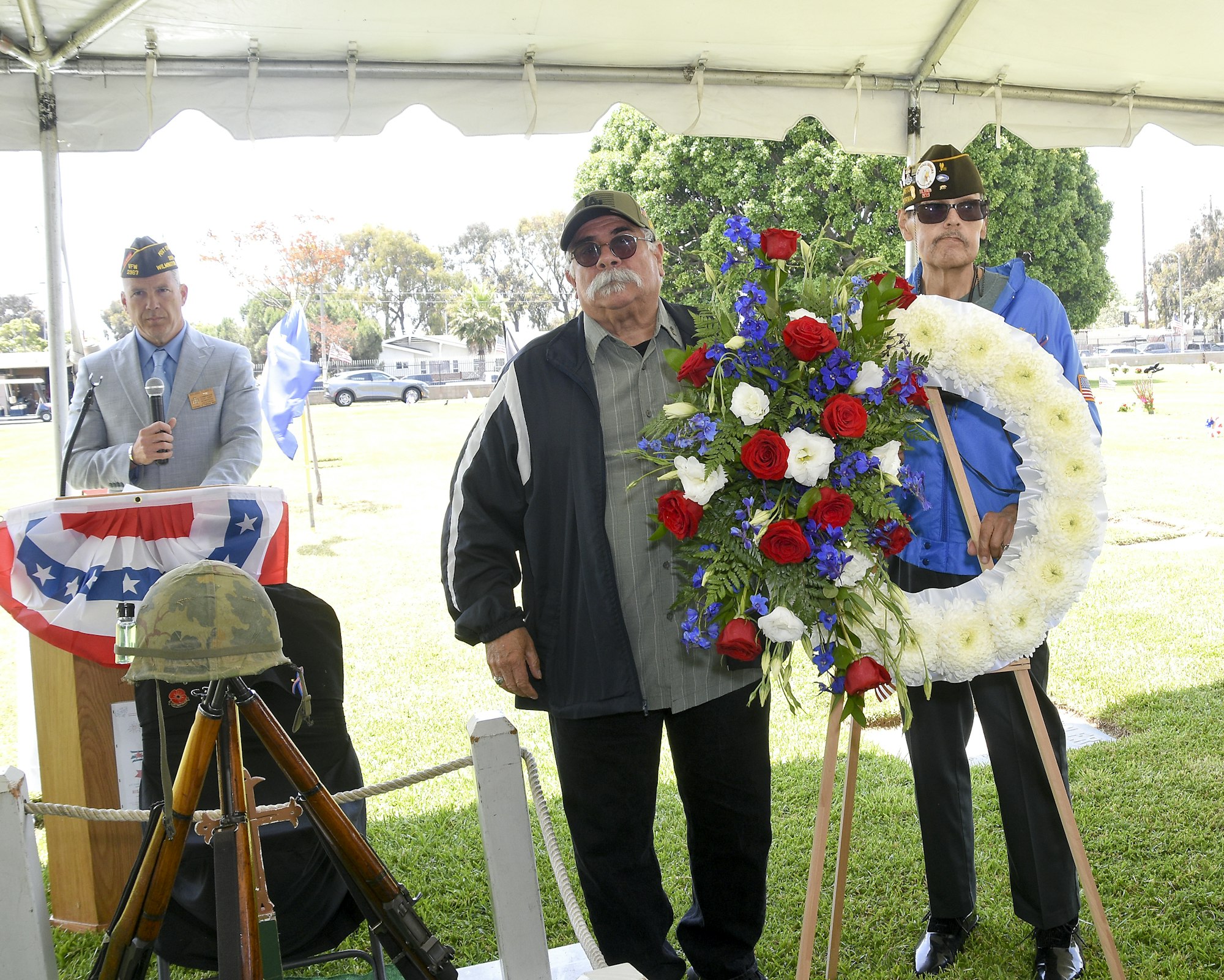 A ceremony with two men holding a floral wreath, a speaker with a microphone, military headgear, and flags under a tent.