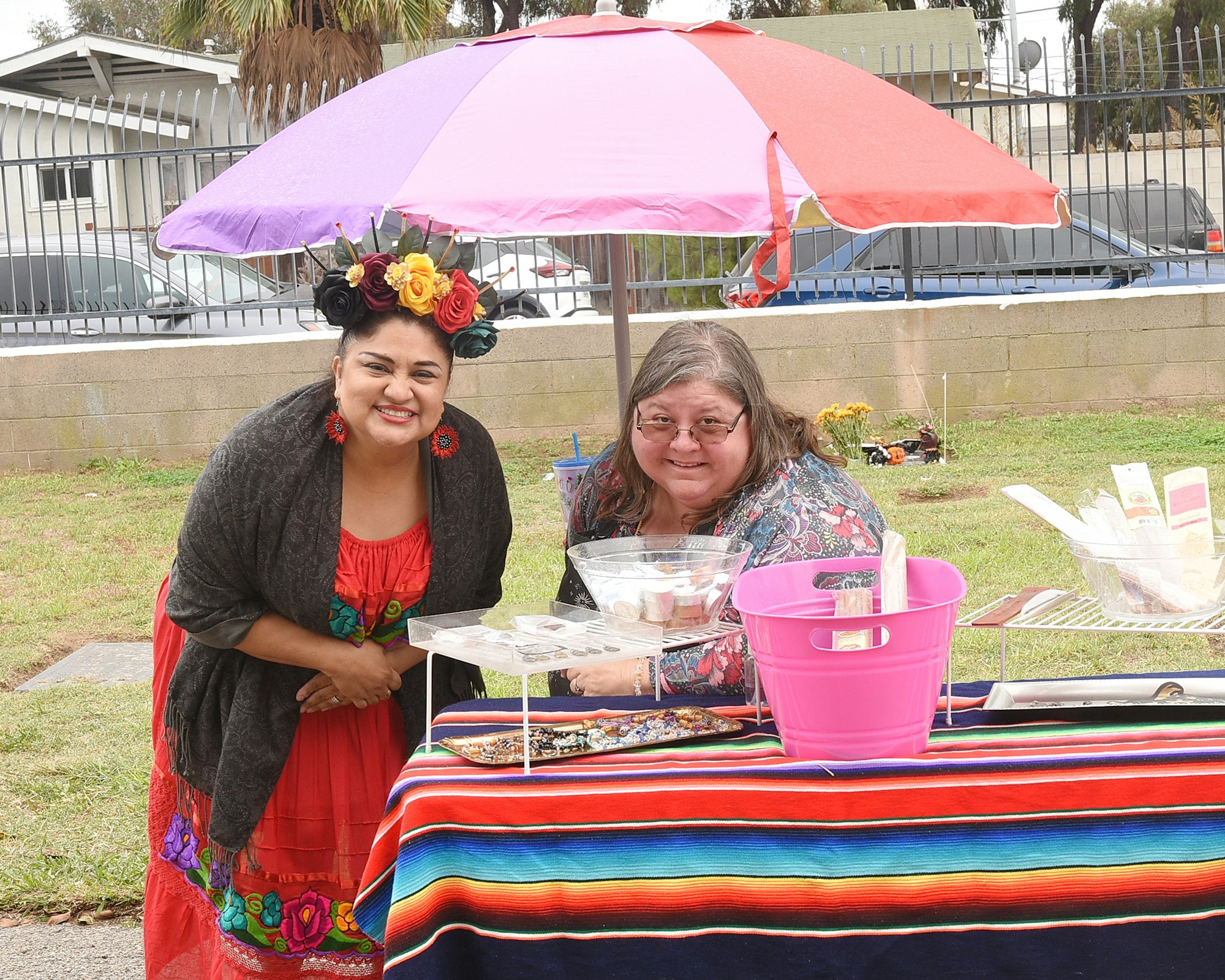 Two smiling women at a colorful outdoor table with food, a multi-colored umbrella overhead.