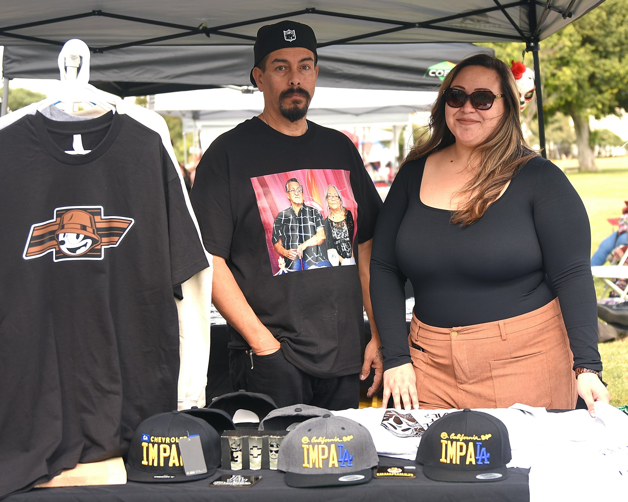 Two people standing behind a table with merchandise like t-shirts and hats at an outdoor event.