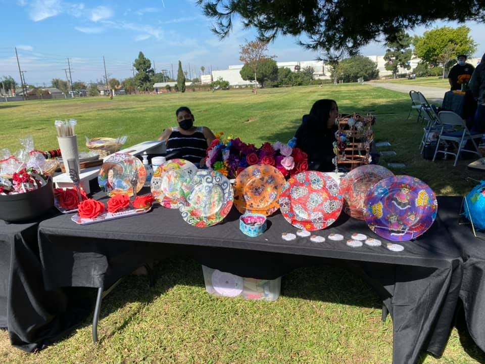 A table with colorful decorative plates, flowers, and crafts at an outdoor market. Two people are seated nearby.