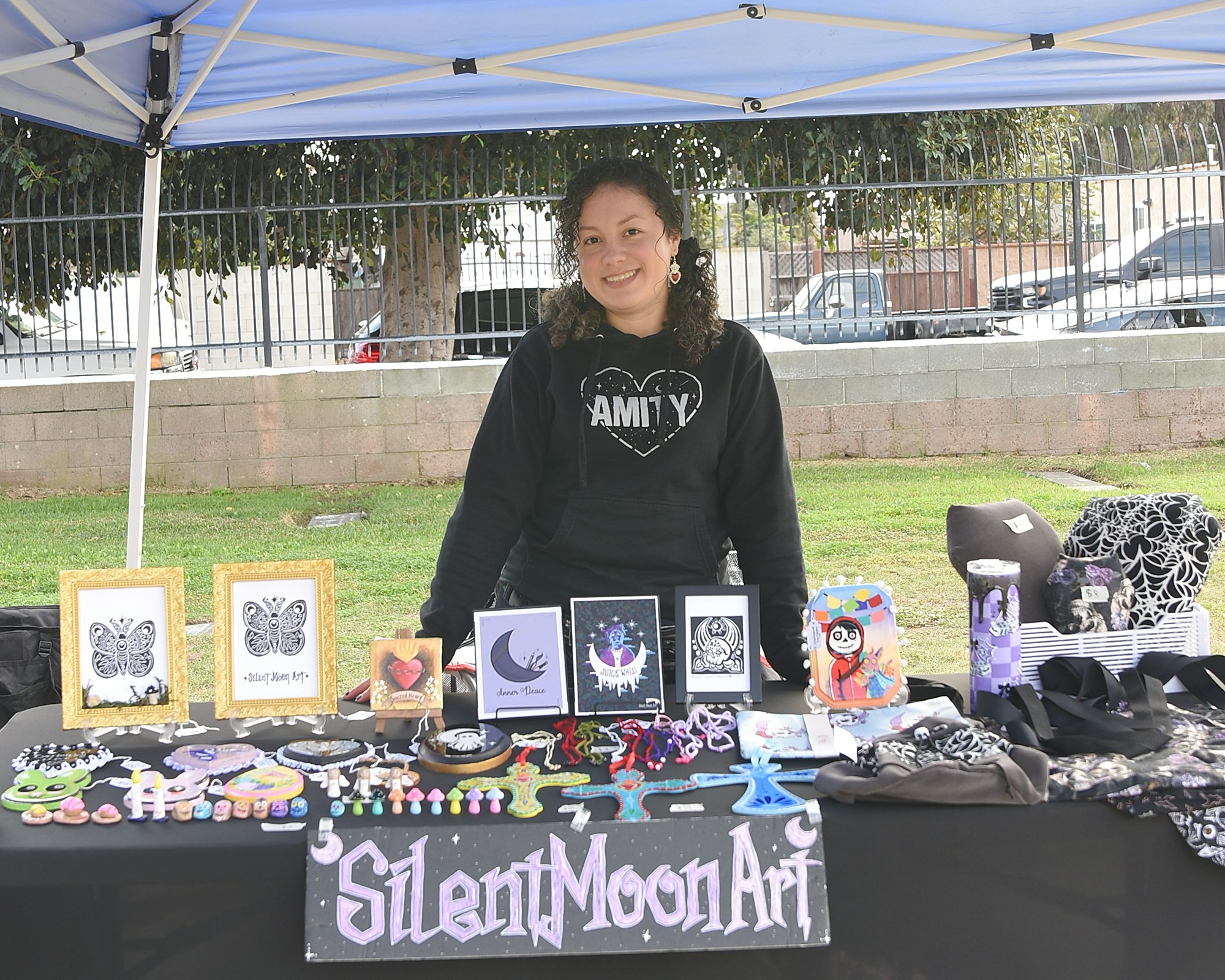 A person stands at a stall displaying artwork and handmade items under a canopy.
