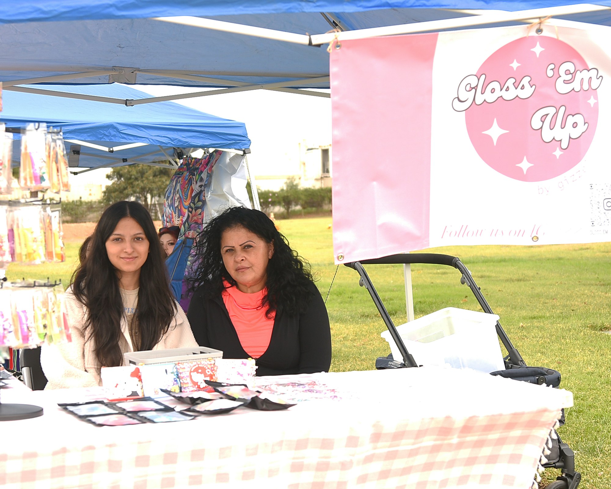 Two women at a market stall with a "Gloss 'Em Up" banner, selling beauty products.
