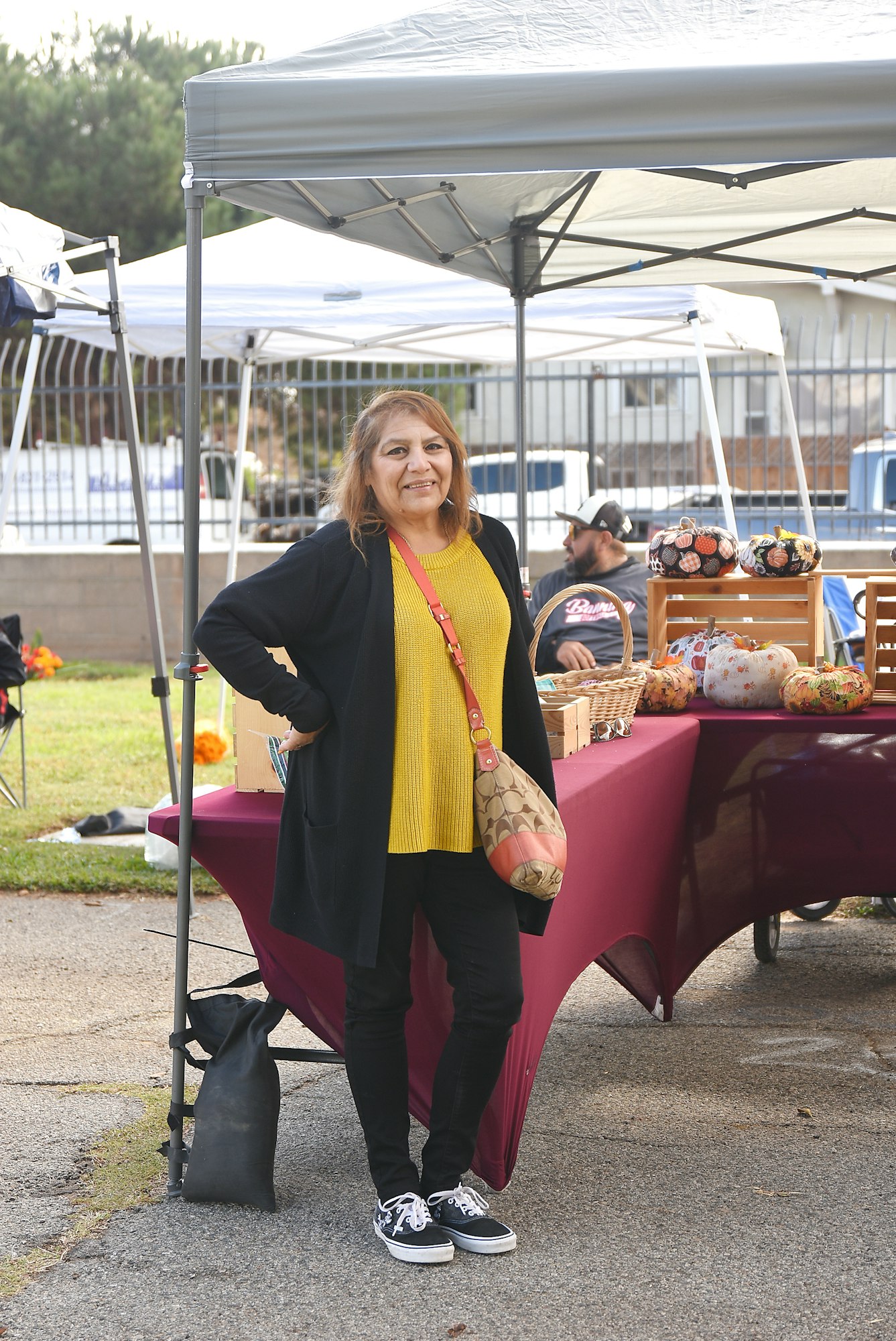 A person standing at a farmers market with tents and craft items in the background.
