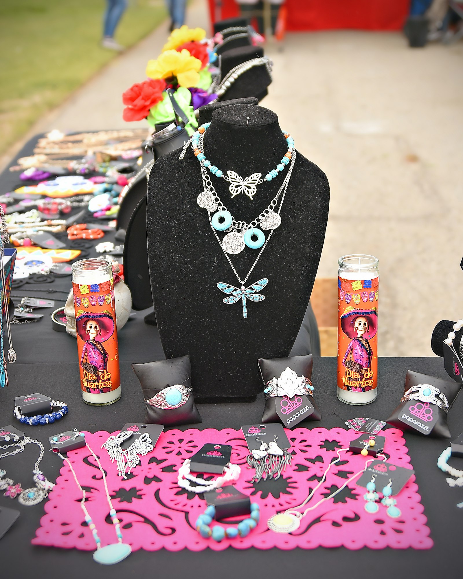 A table displays a variety of colorful jewelry, with necklace busts and decorative candles, likely at a crafts market.