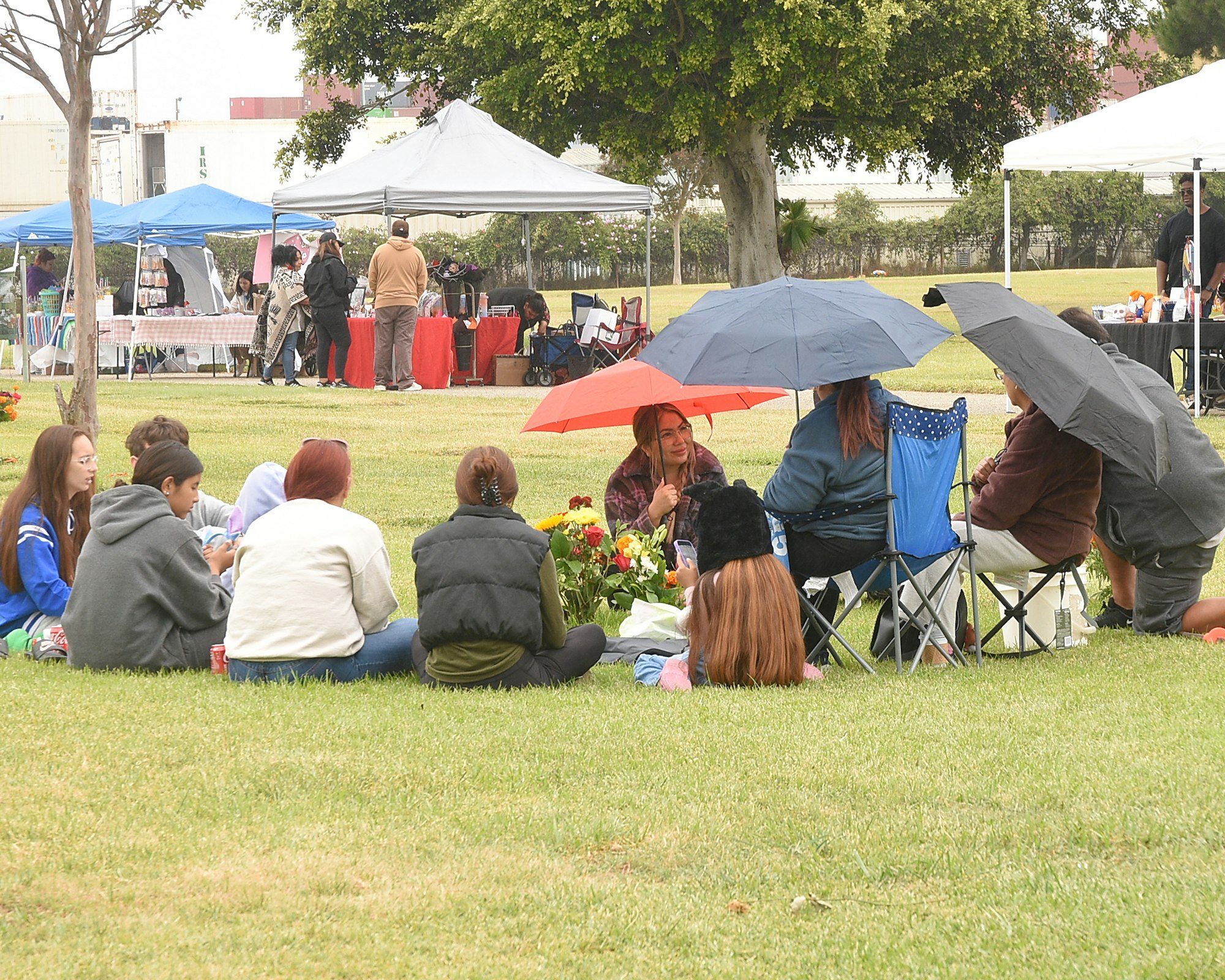 People gathered in a park, some under umbrellas, with tents in the background.
