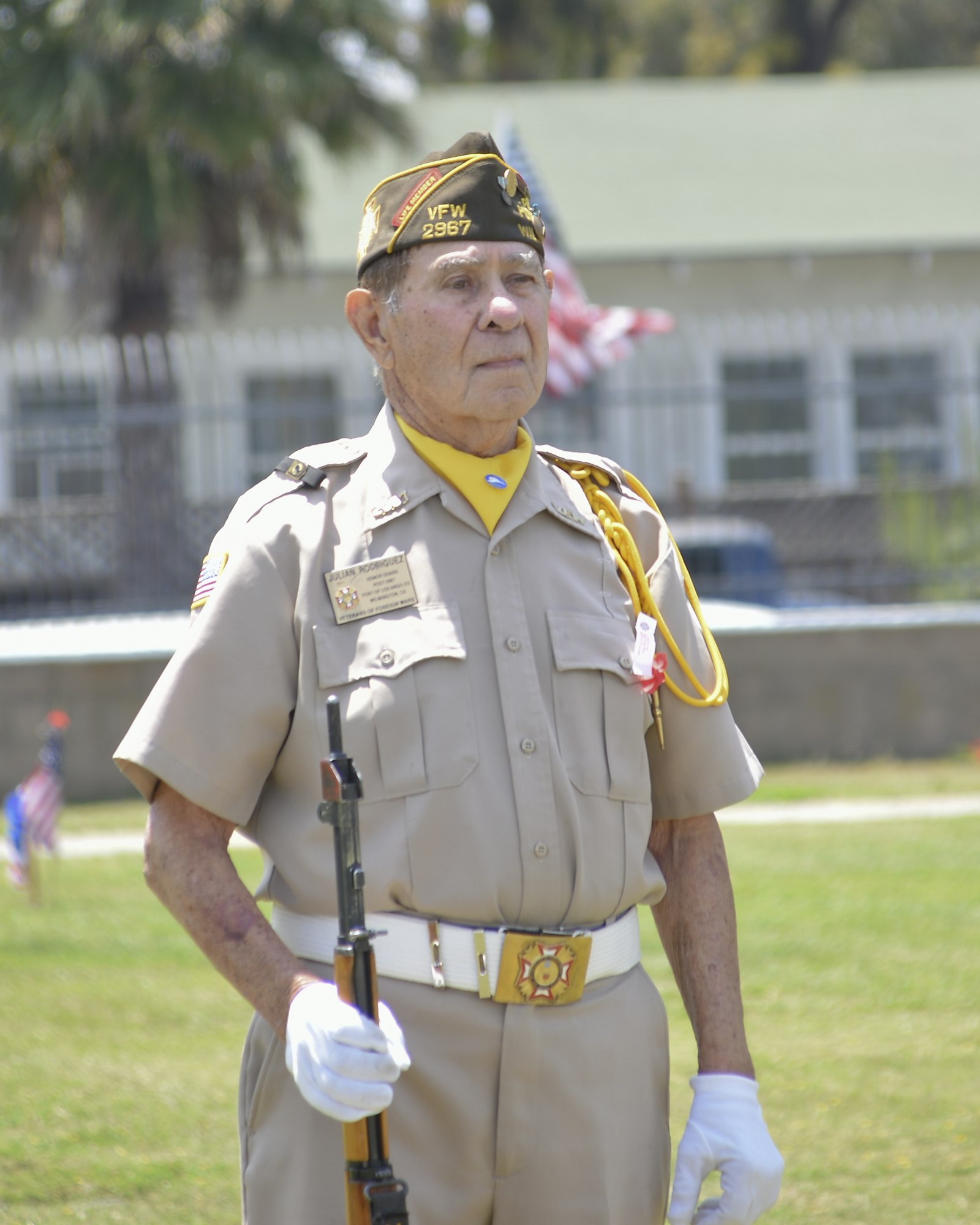 An elderly veteran in uniform with a cap and medals, holding a rifle, at an outdoor event.
