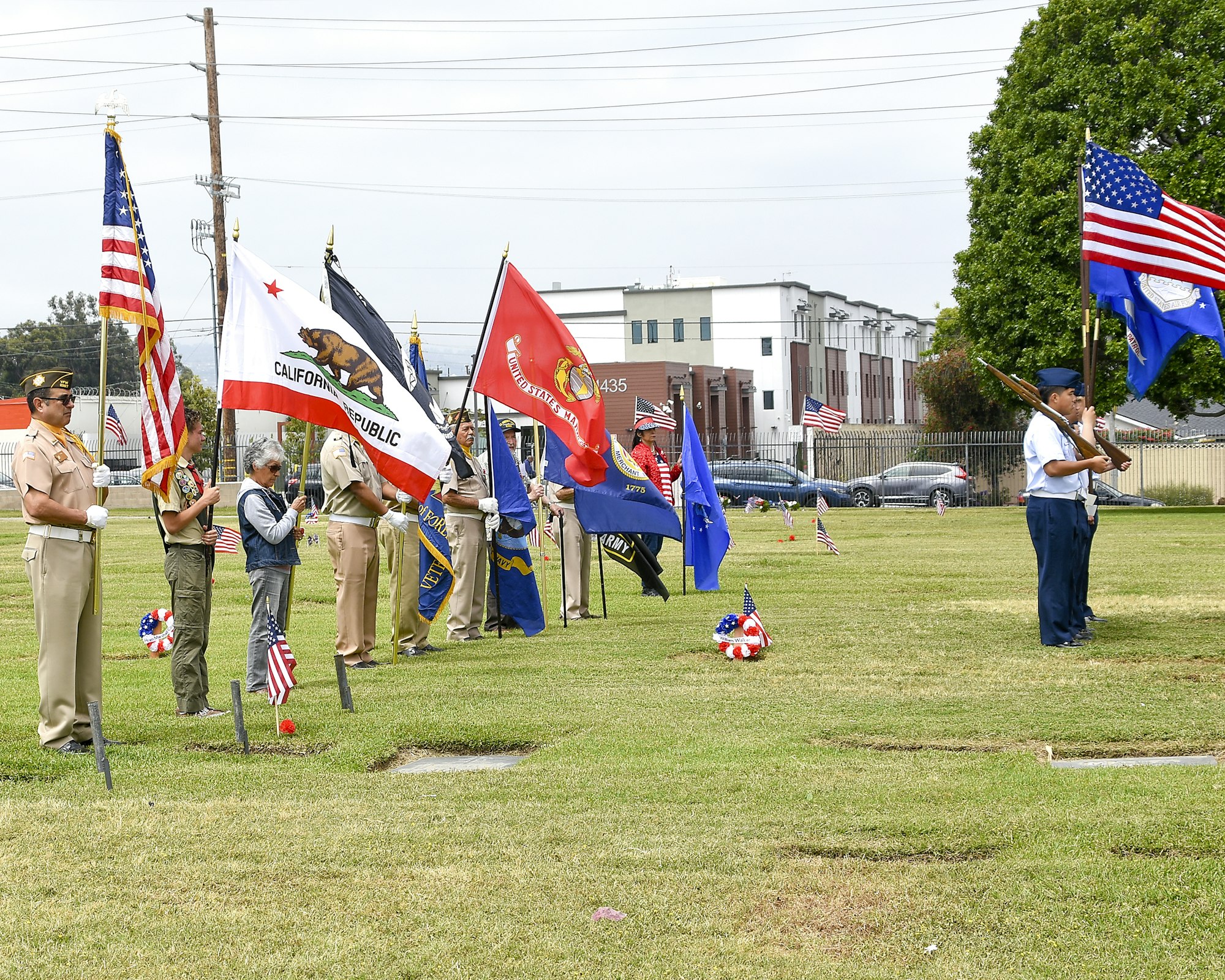 People holding various flags, including the US and California flags, in a grassy area, likely during a ceremonial event.