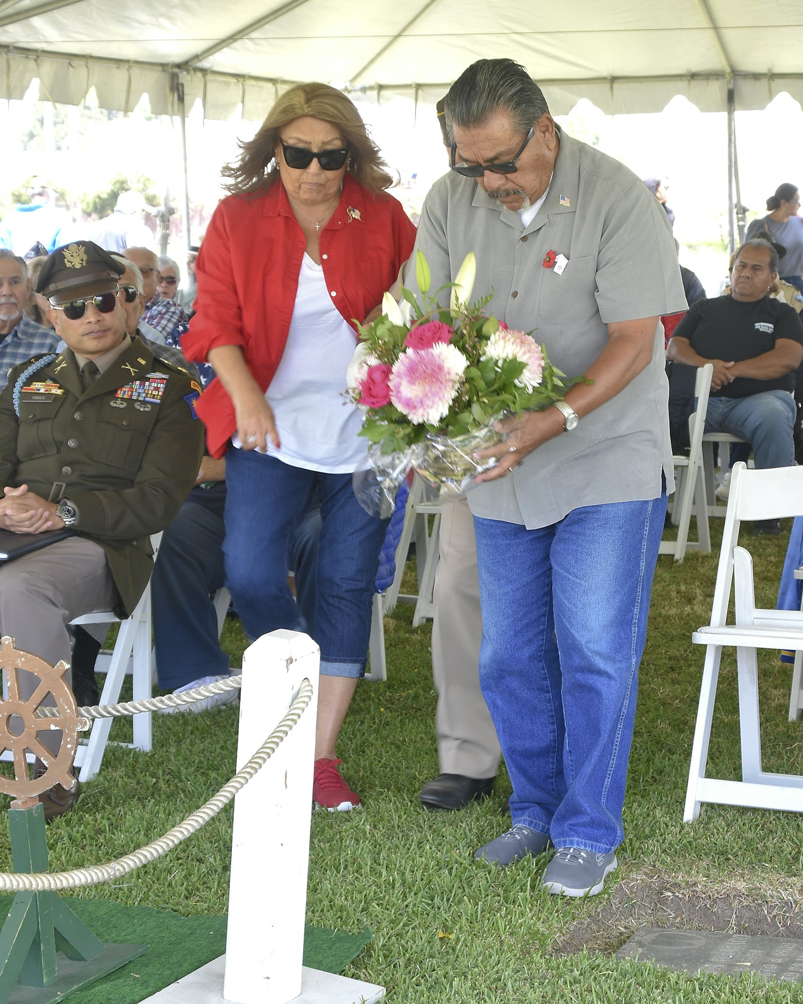 Two individuals are placing flowers near a headstone, under a tent with seated attendees, possibly at a commemoration event.