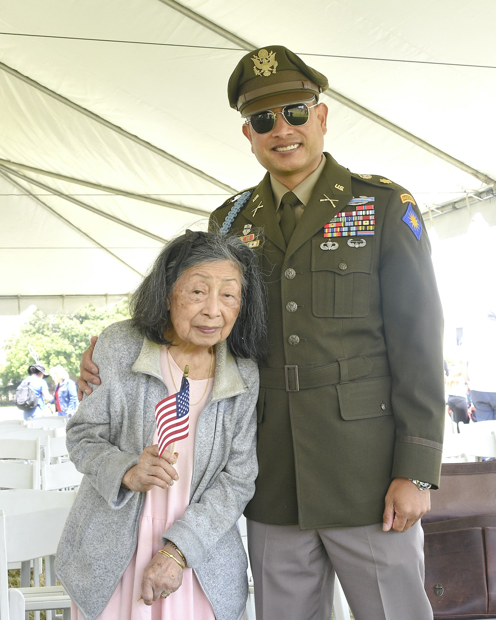An elderly woman with an American flag and a man in military uniform standing side by side, smiling.