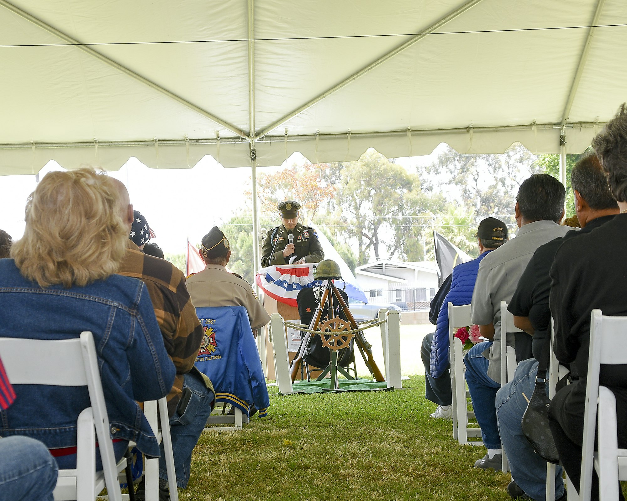 A group listens to a uniformed speaker at an outdoor event under a tent, with a folded American flag in the foreground.