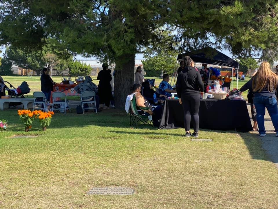 A park scene with people gathered around tables, enjoying camaraderie and activities, under trees and a clear sky.