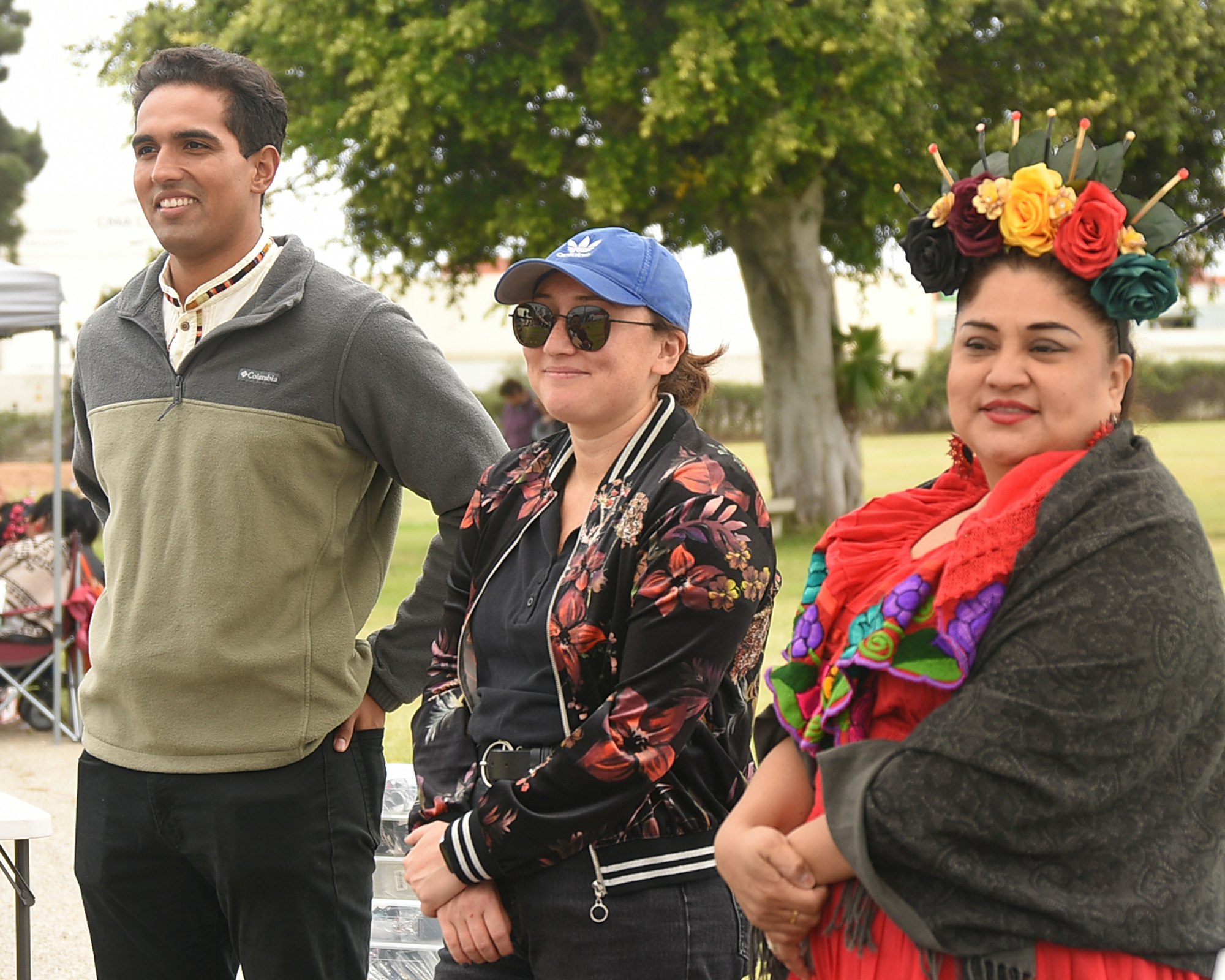 Three people standing outdoors; one in vibrant traditional Mexican attire with a flower crown.