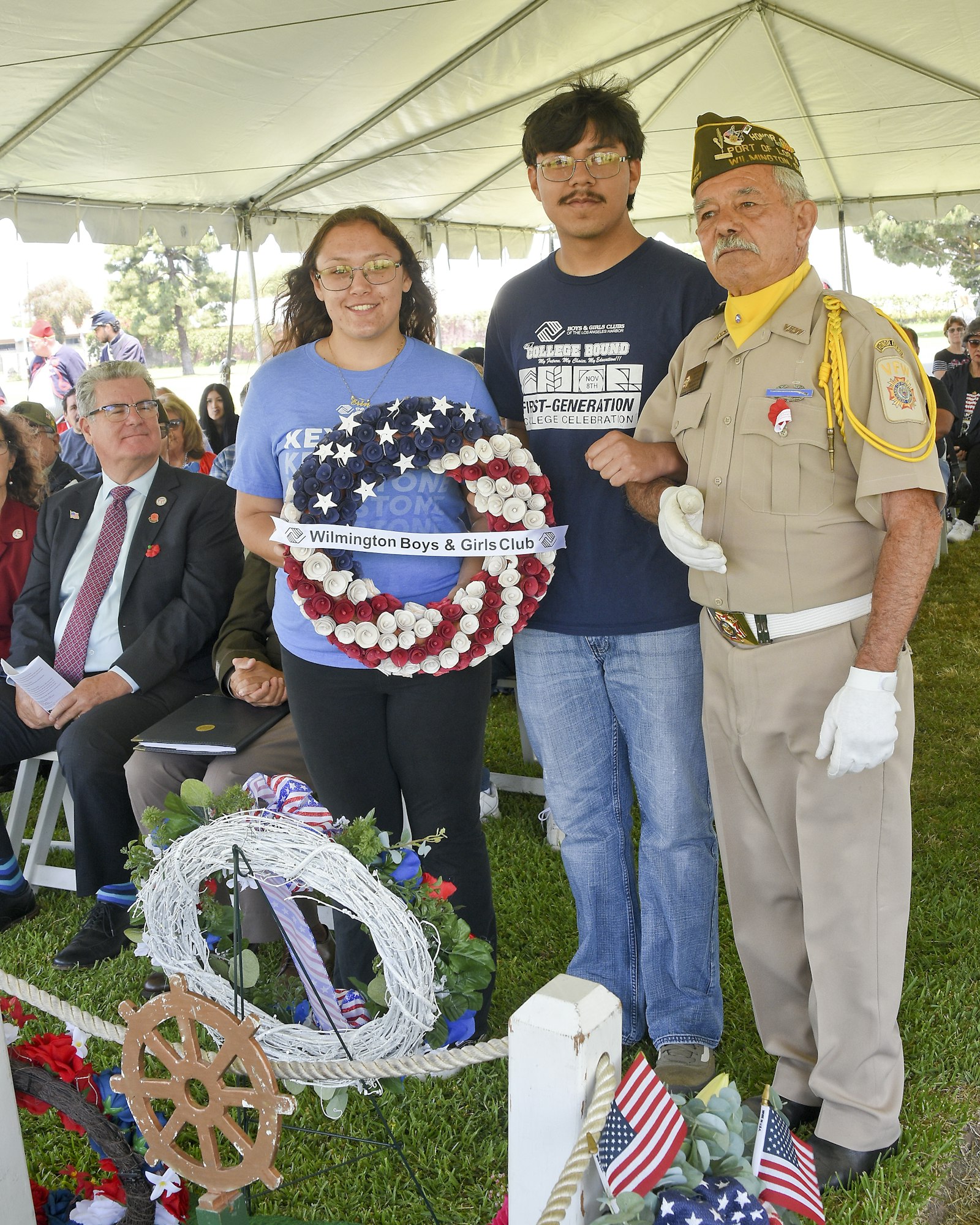 Three people pose with a star-spangled wreath, under a tent, at an outdoor event, possibly patriotic, with onlookers seated in the background.