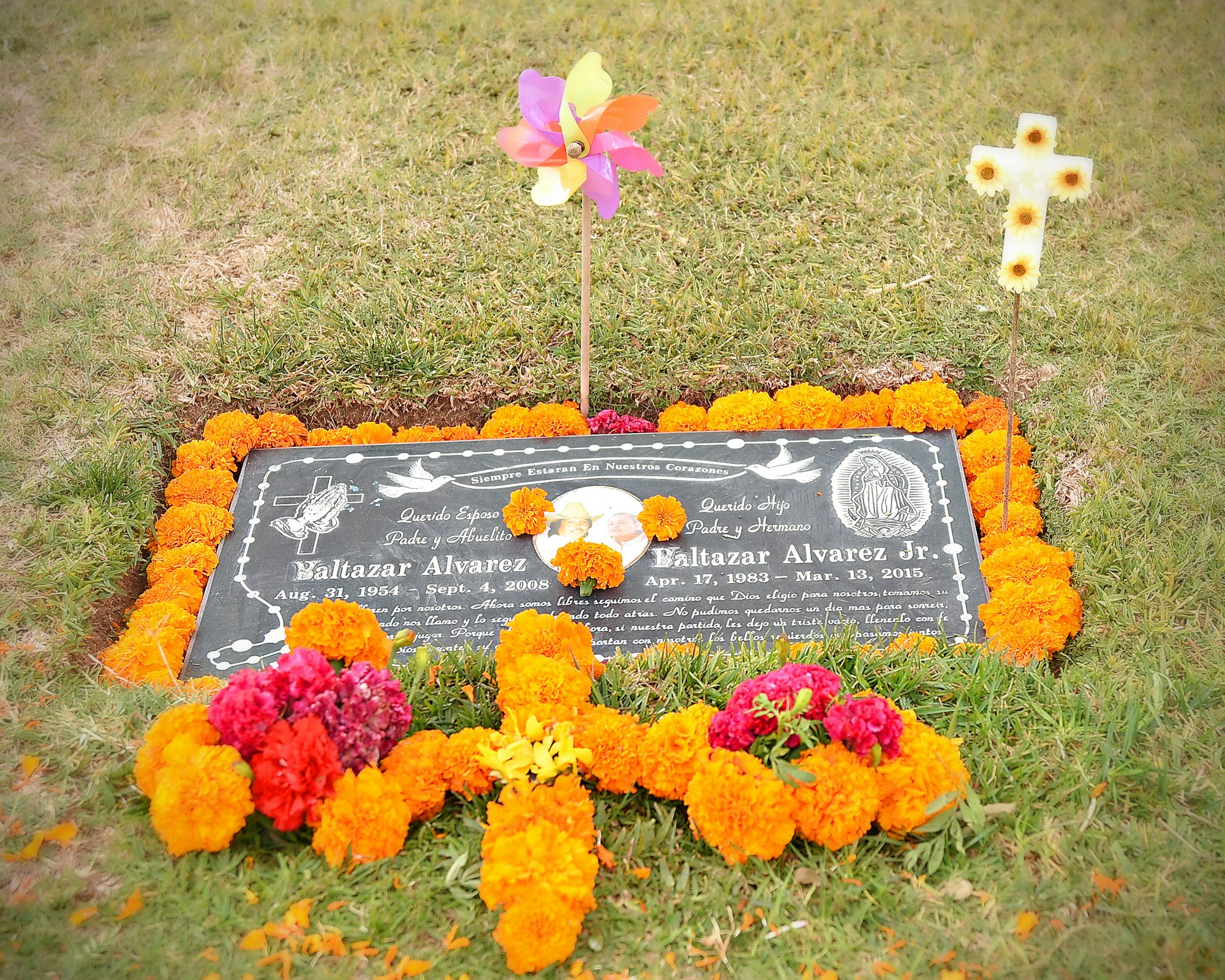 Grave adorned with flowers, a pinwheel, and a cross, possibly for Día de los Muertos celebration.