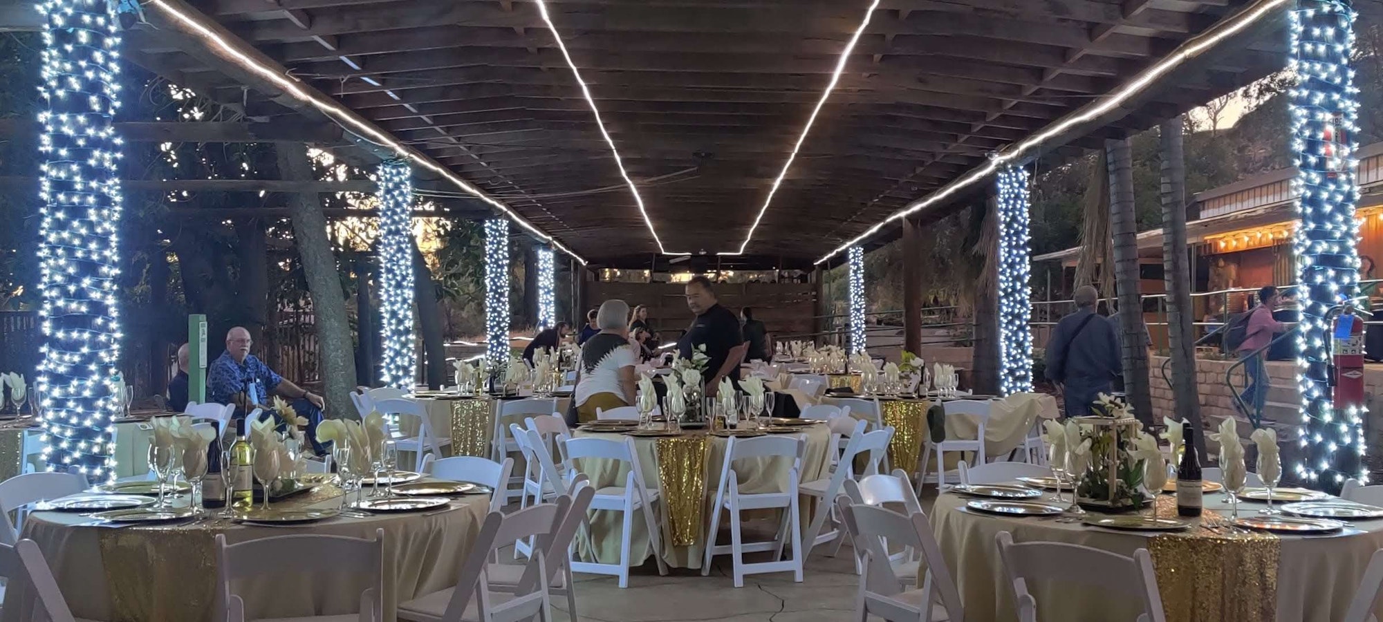 Outdoor event setup with tables, chairs, and white lights wrapped around columns under a wooden pergola.