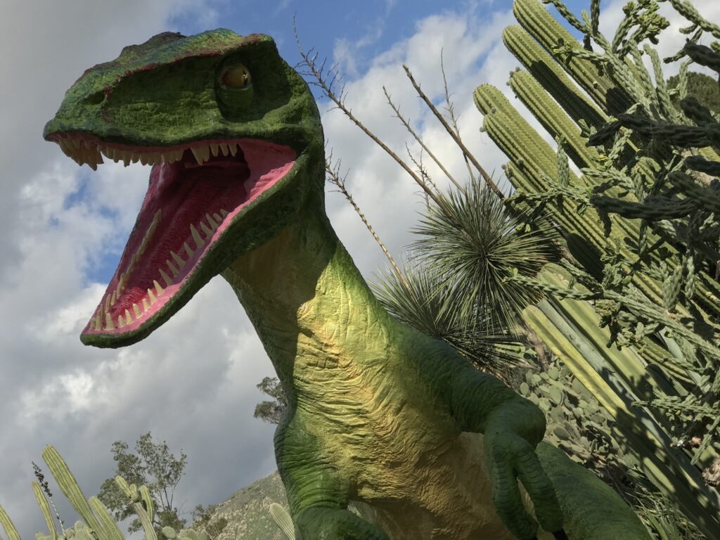 A green dinosaur statue with an open mouth, surrounded by cacti under a partly cloudy sky.