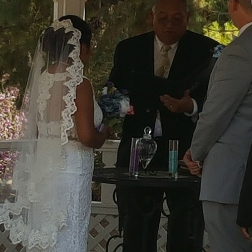 A wedding ceremony with a couple facing an officiant, a table with sand vases, and the bride in a white dress with a veil.