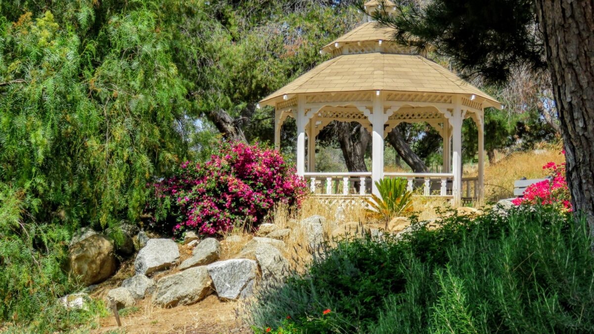 A white gazebo surrounded by trees, rocks, and colorful flowers in a garden setting.
