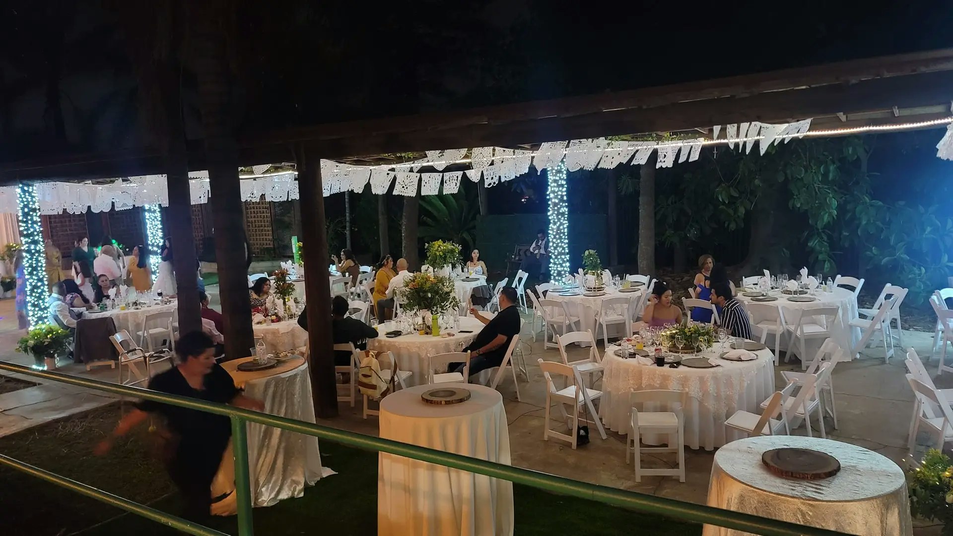 An outdoor event with decorated tables, festive lights, and people mingling and seated at round tables.