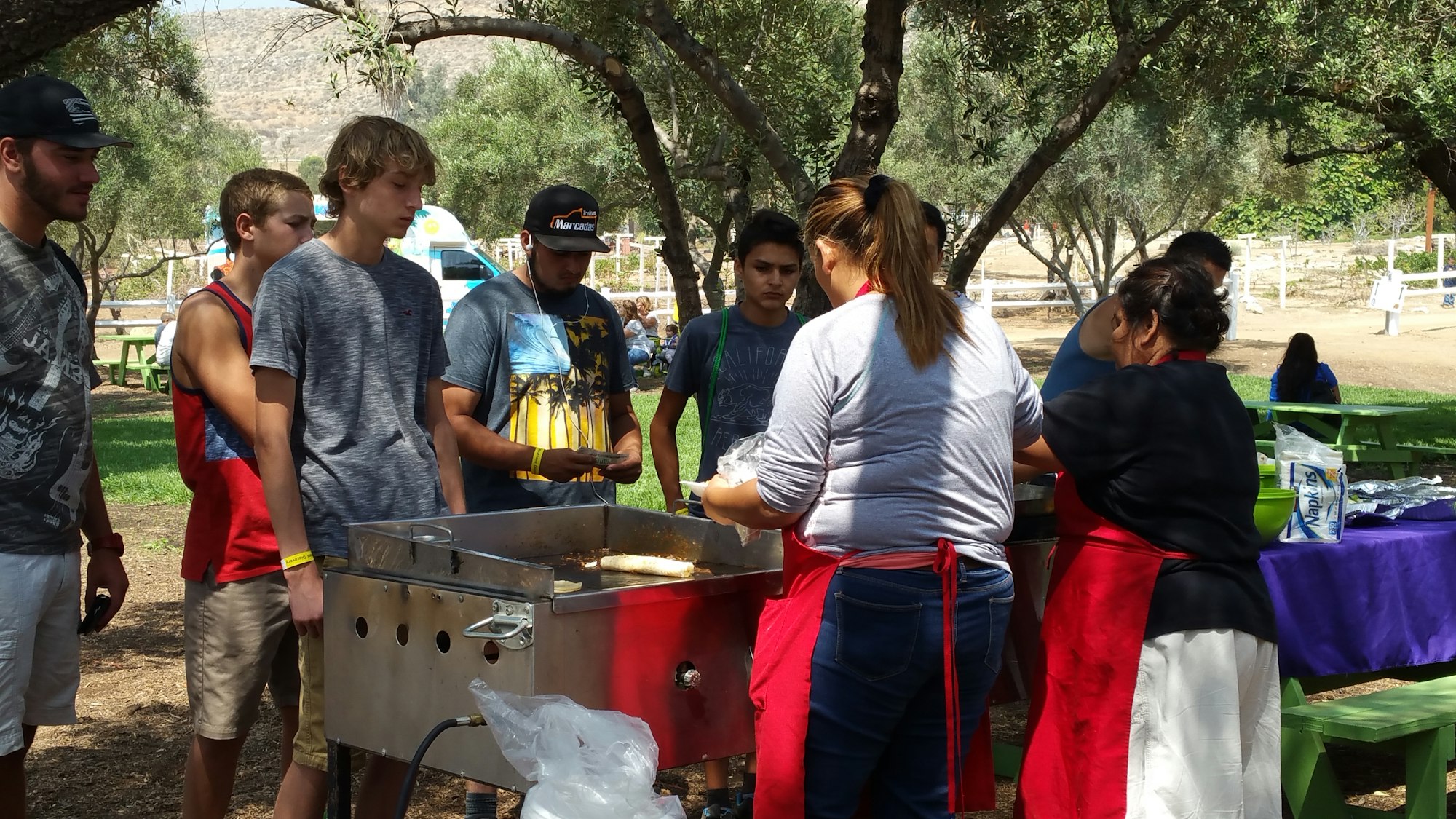 People gathered outdoors around a cooking station, some in aprons, others waiting. Trees and picnic areas in the background.