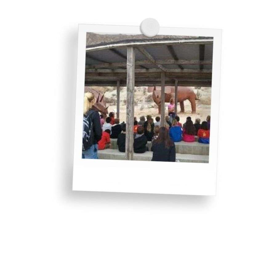 A group of people under a shelter watching a large animal sculpture outdoors.
