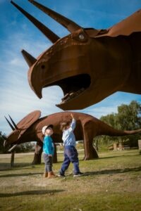 Two kids stand near large metal dinosaur sculptures in a park.