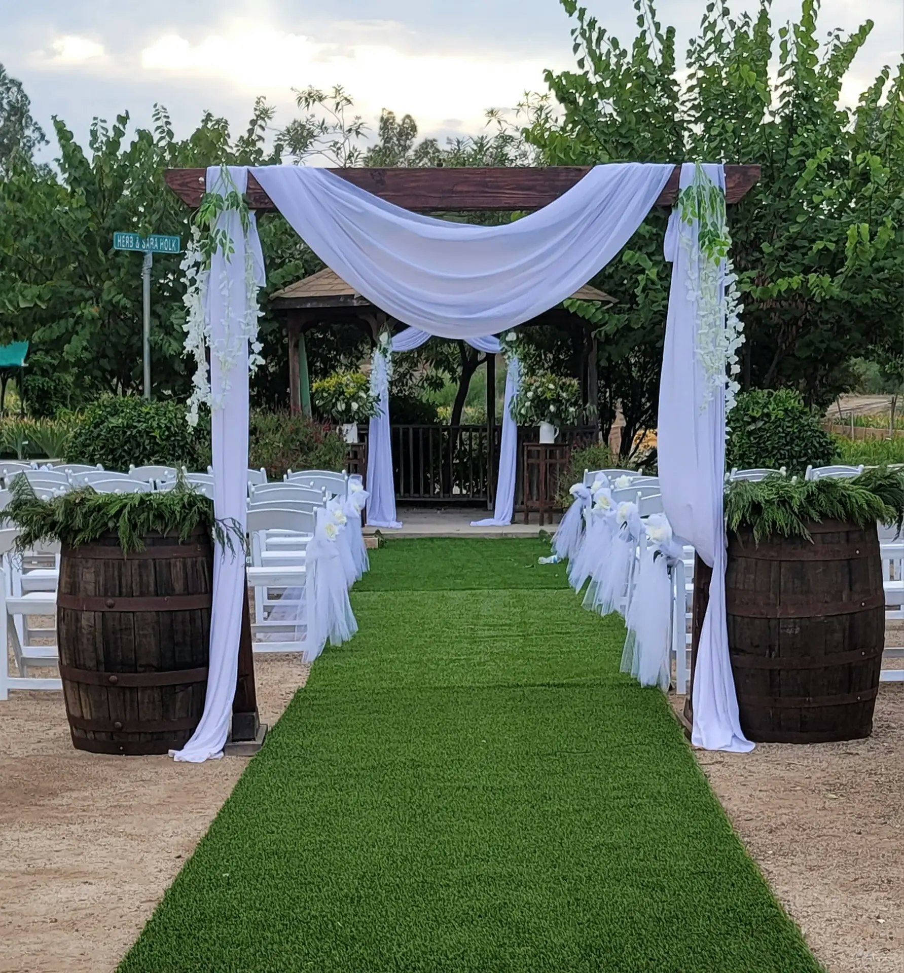 Outdoor wedding setup with an aisle, white chairs, draped archway, and greenery.