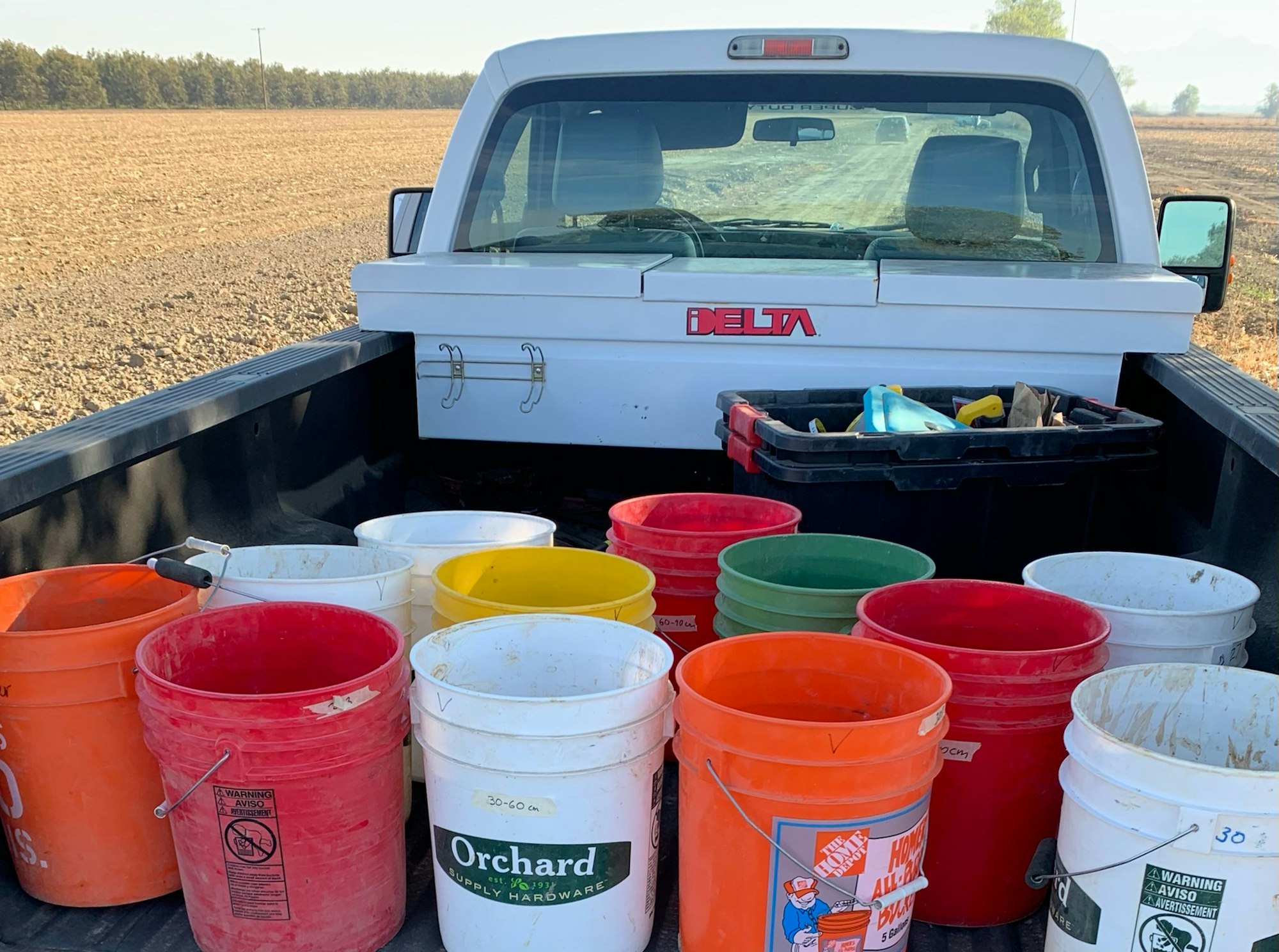 The image shows the back of a pickup truck with several colorful buckets and a toolbox, set against an agricultural field backdrop.