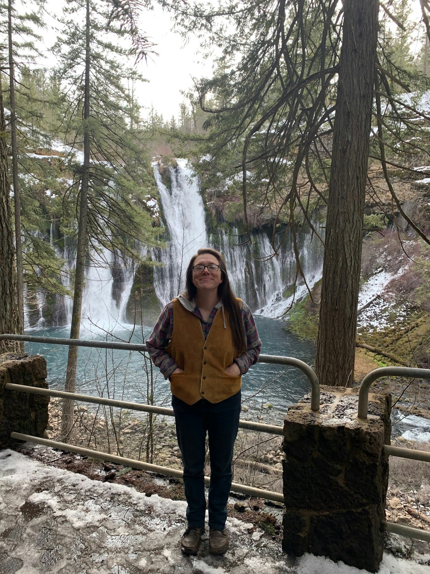A person stands smiling in front of a waterfall, surrounded by trees in a natural setting, with their hands in their vest pockets wearing boots and jeans.