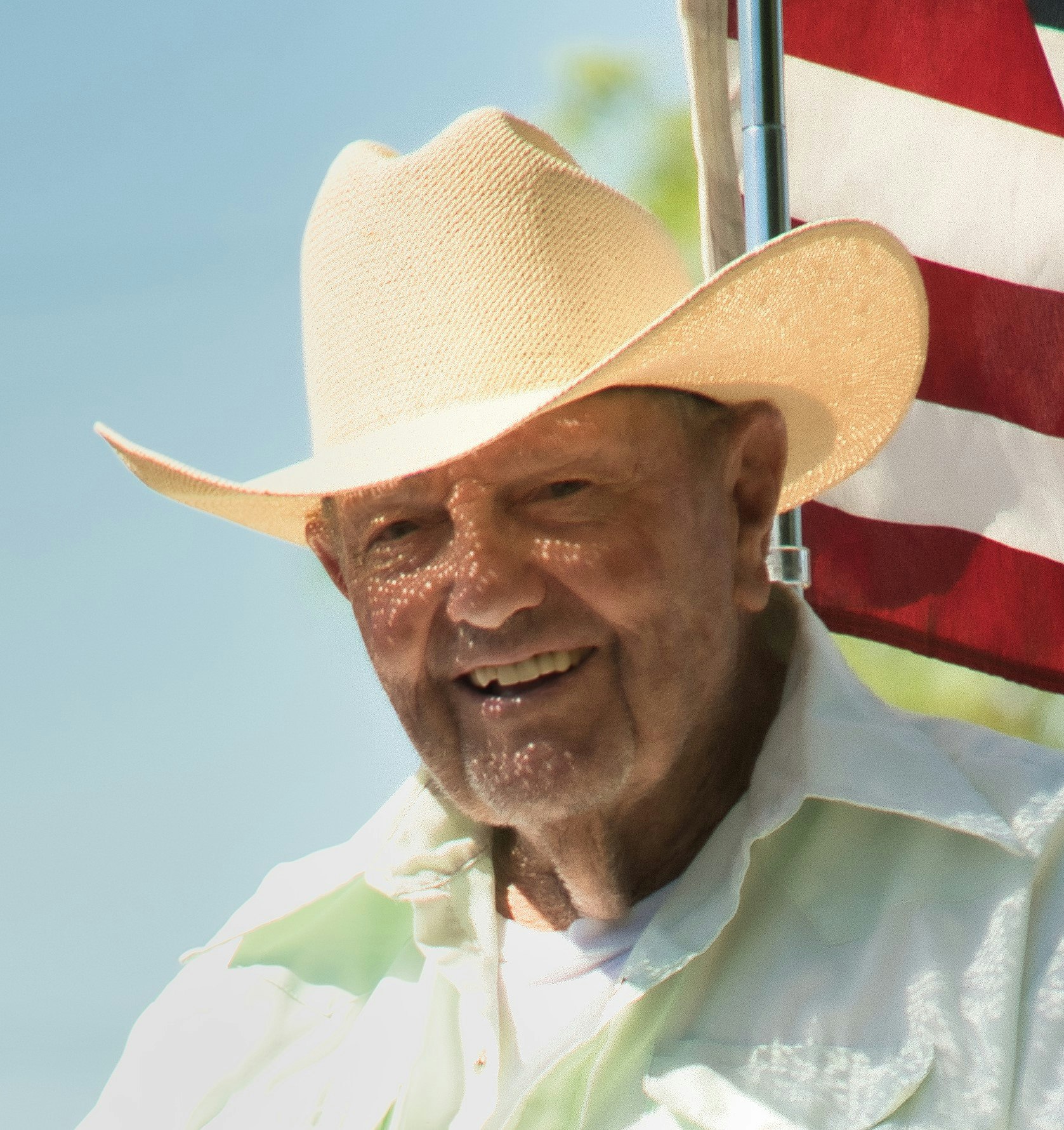 An elderly man with a smiling face wearing a straw hat, standing in front of an American flag. The background is bright and cheerful.