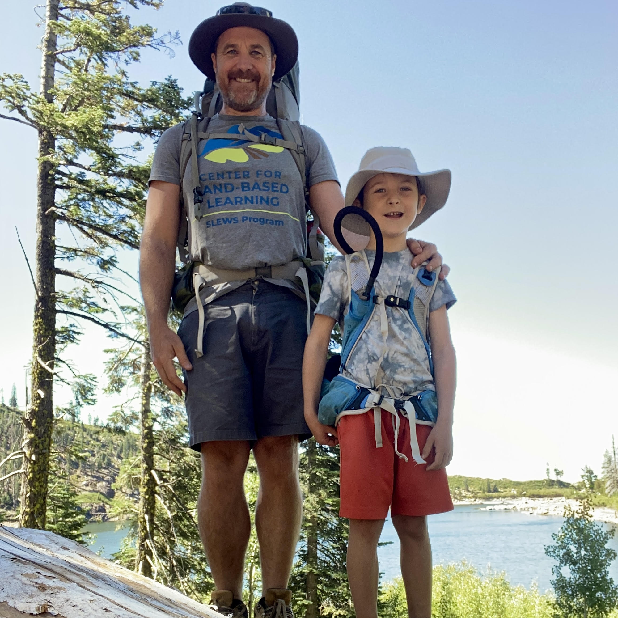 A man and a child stand outdoors, both wearing hats and backpacks, surrounded by trees with a lake in the background. They smile for the photo.