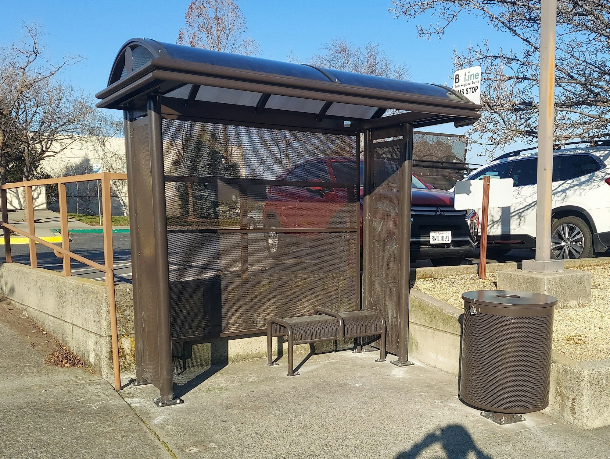 A bus stop with a shelter, bench, trash can, and parked cars nearby.