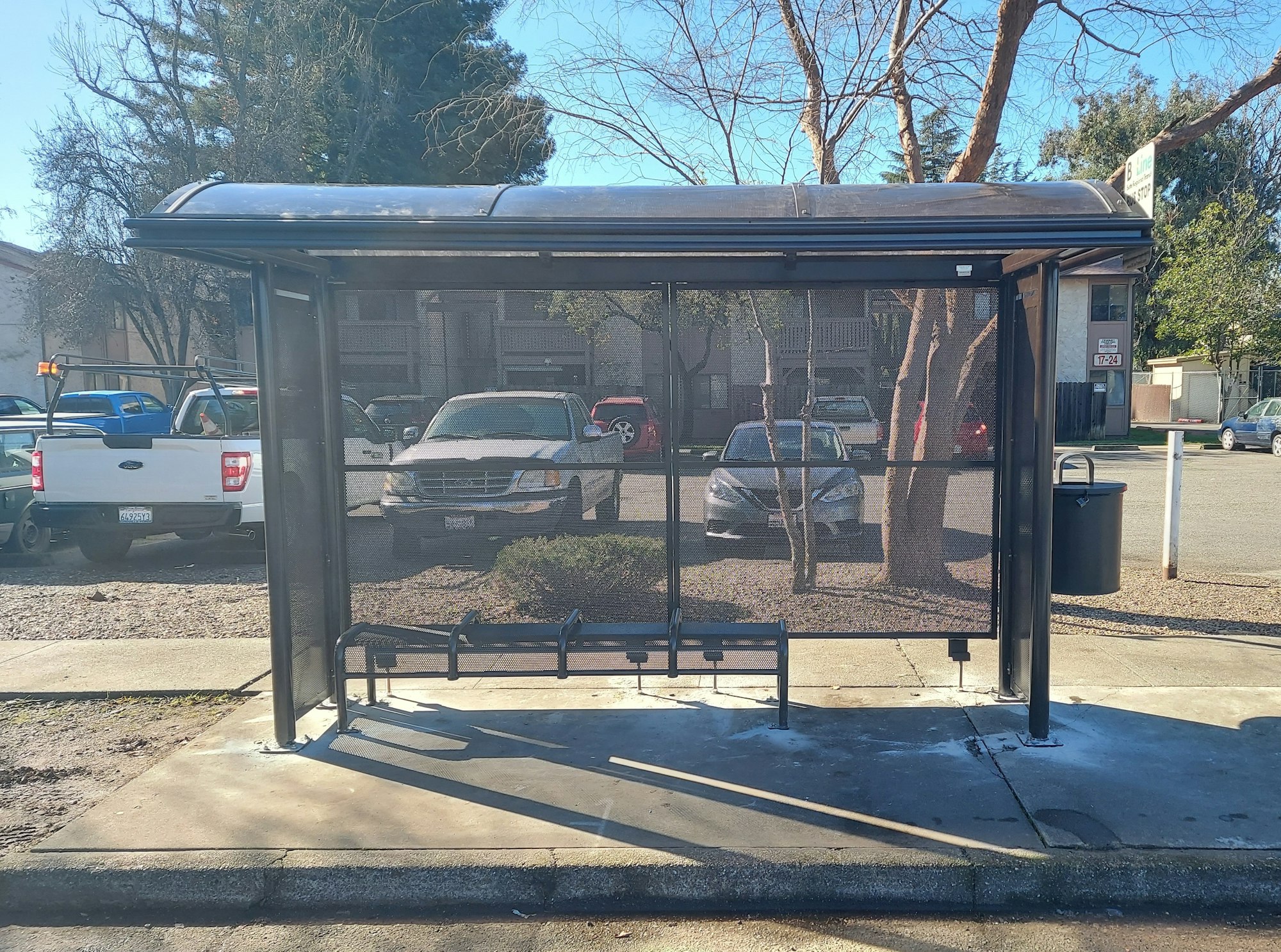 A bus stop shelter with a bench and trash can, surrounded by parked cars and trees.
