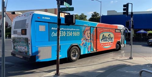 A bus with an "AttaBoy Plumbing" advertisement, parked near a street intersection.