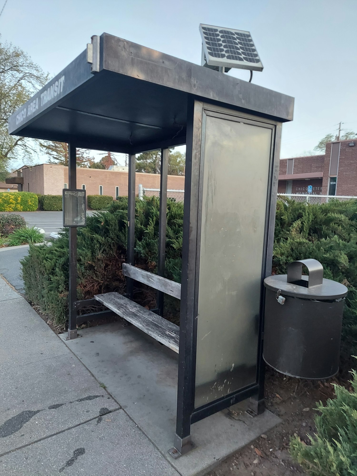 A small bus stop shelter with a bench, solar panel on top, and a trash can next to it.