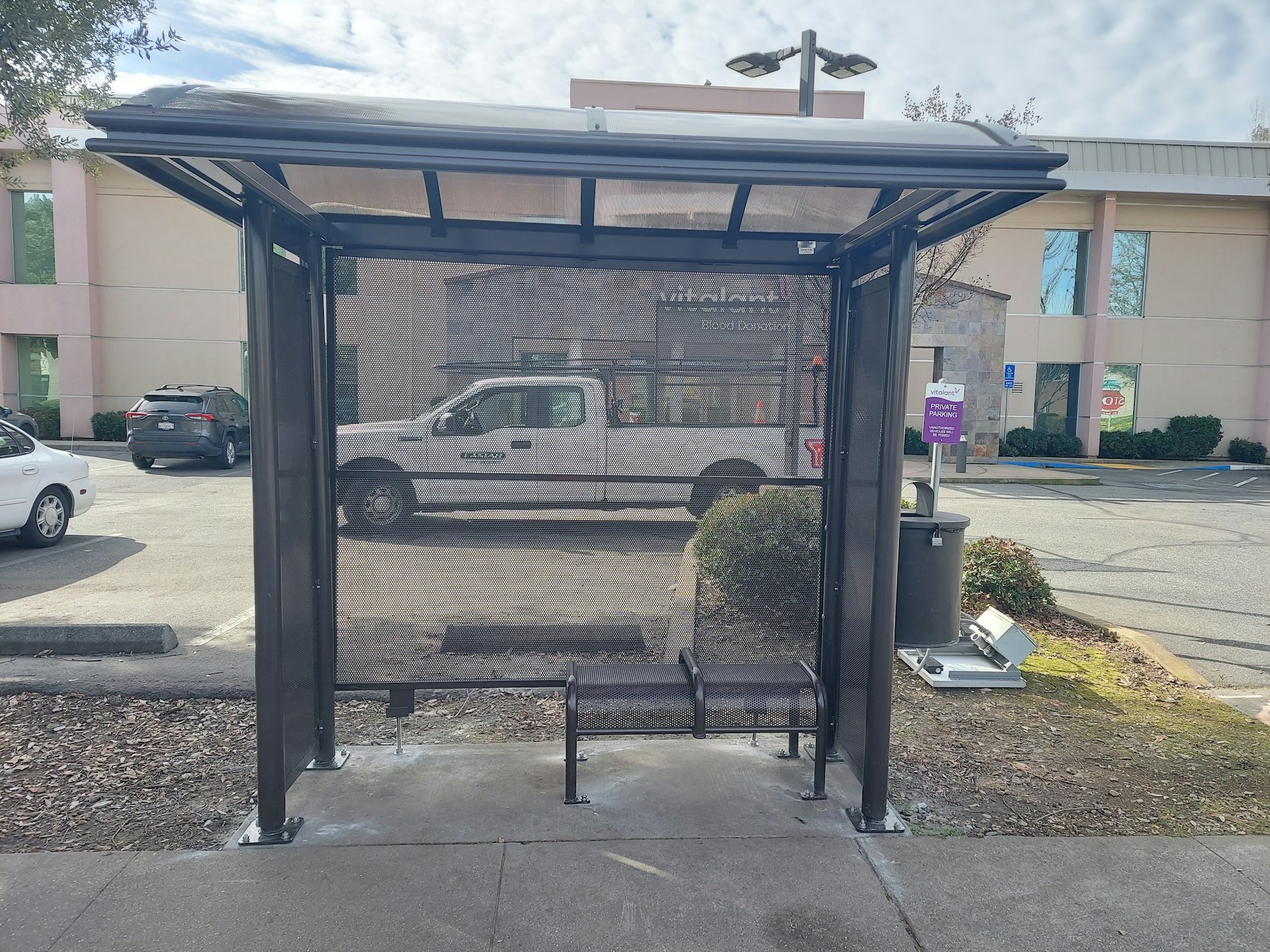 Bus stop shelter with a bench, located in a parking lot near a building.
