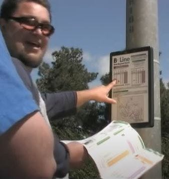 A person points at a bus schedule on a pole, holding a map in hand.