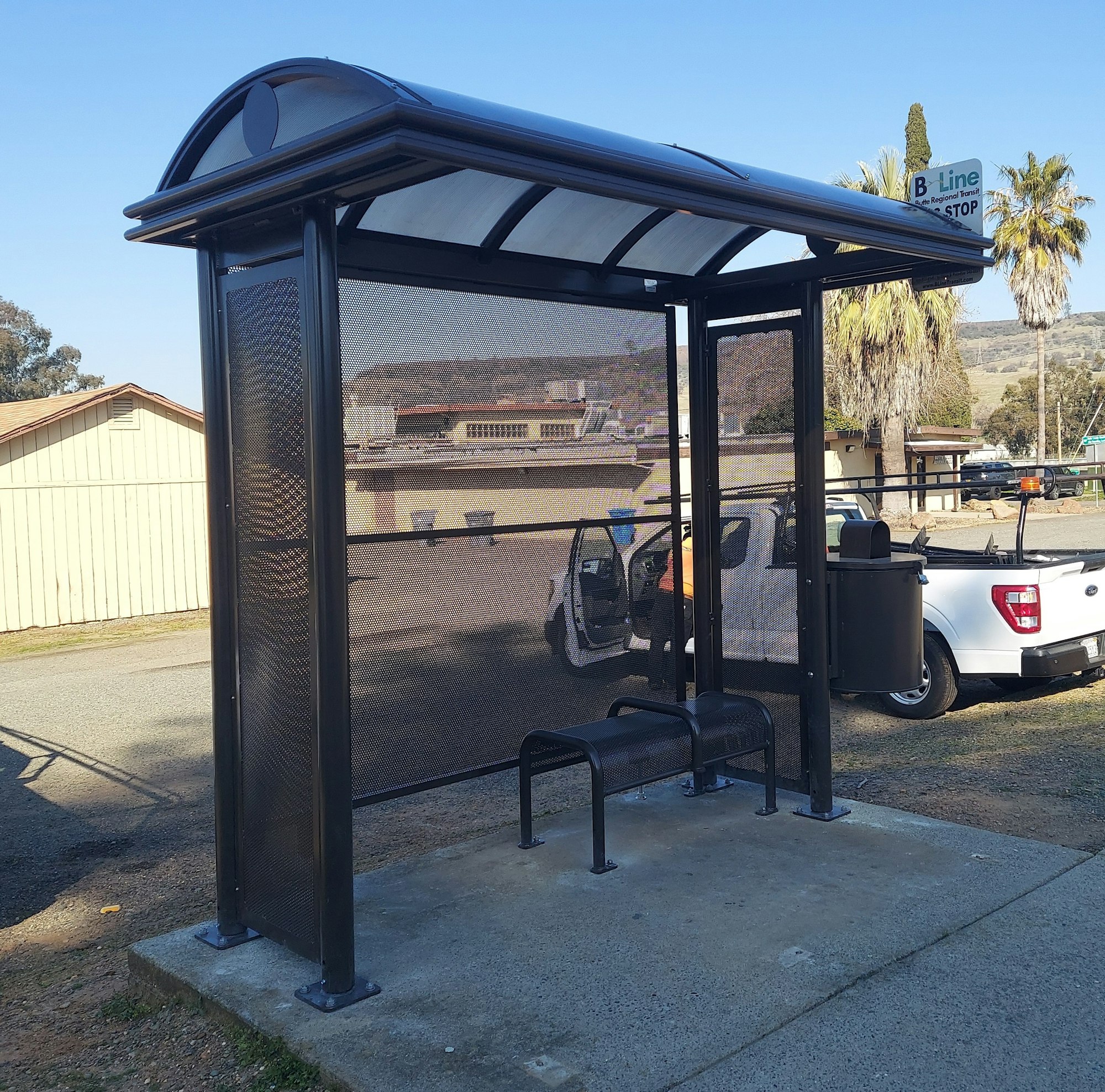 A bus stop shelter with a bench and a trash can, next to a white truck parked on the street.