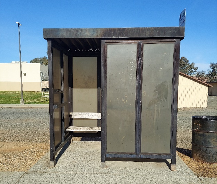 A small, weathered bus shelter with a bench, located on a street with a pole and some buildings in the background.
