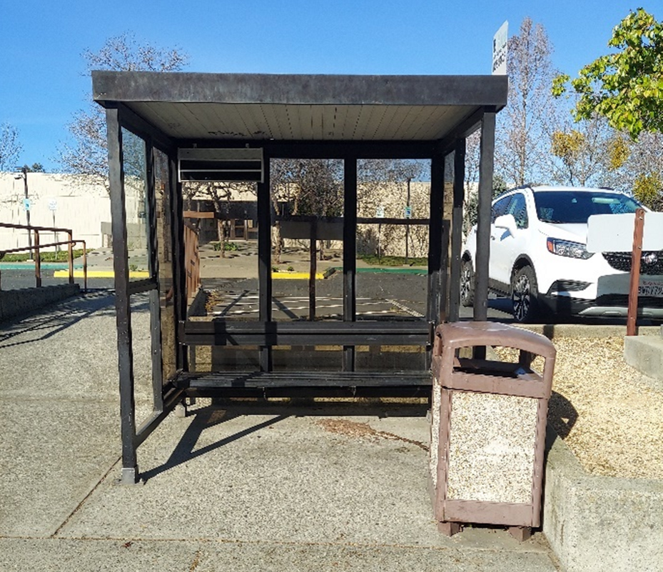 A bus stop shelter with a bench, trash can, and a white car parked nearby under a clear blue sky.