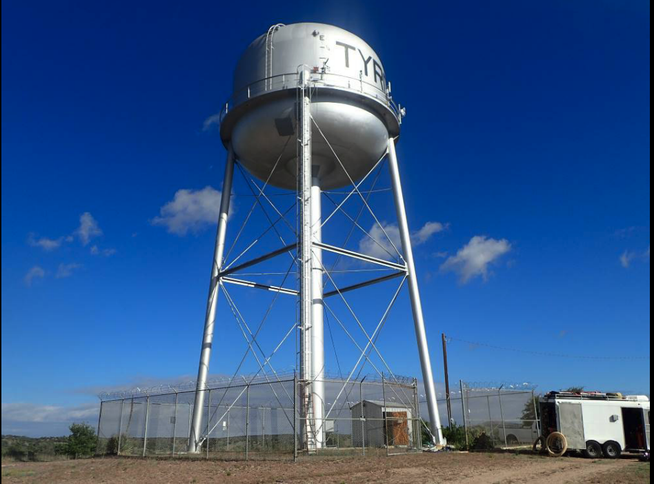 A water tower against a blue sky, surrounded by a fence, with a parked trailer nearby.