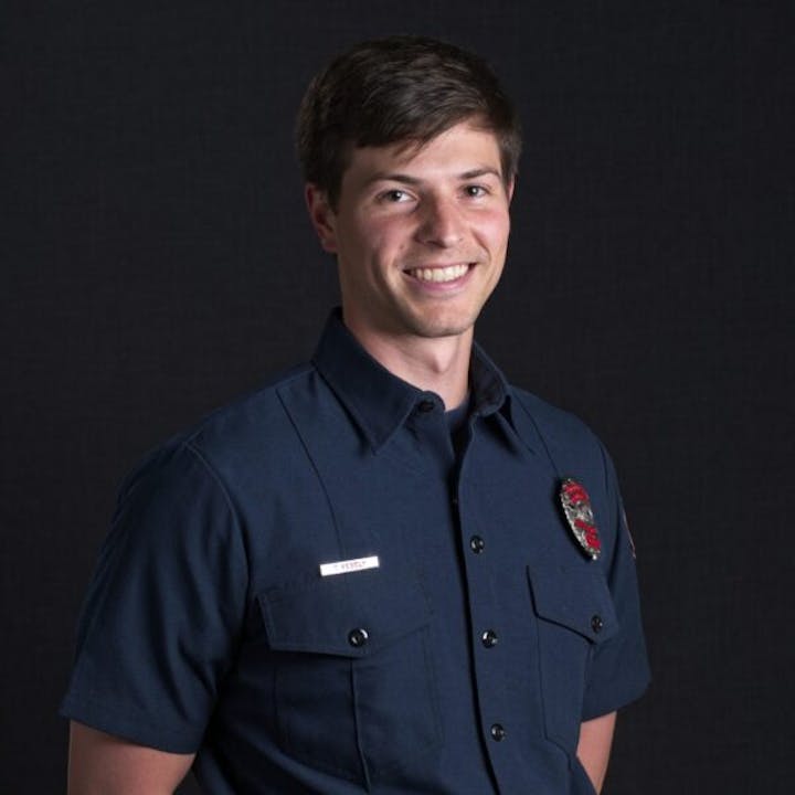 A person in a blue uniform shirt with a badge, smiling, against a dark background.