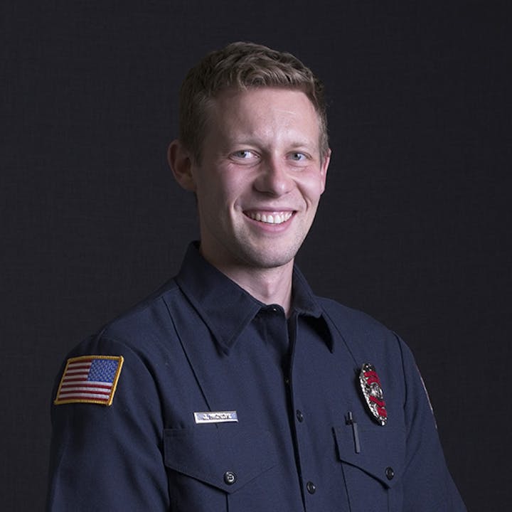 A person in a dark uniform with a badge, smiling against a dark background.